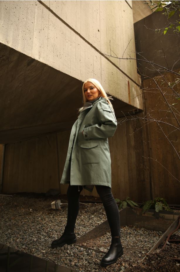 Woman wearing a green unisex parka jacket with utility pockets, and hood patch pockets over a black outfit.