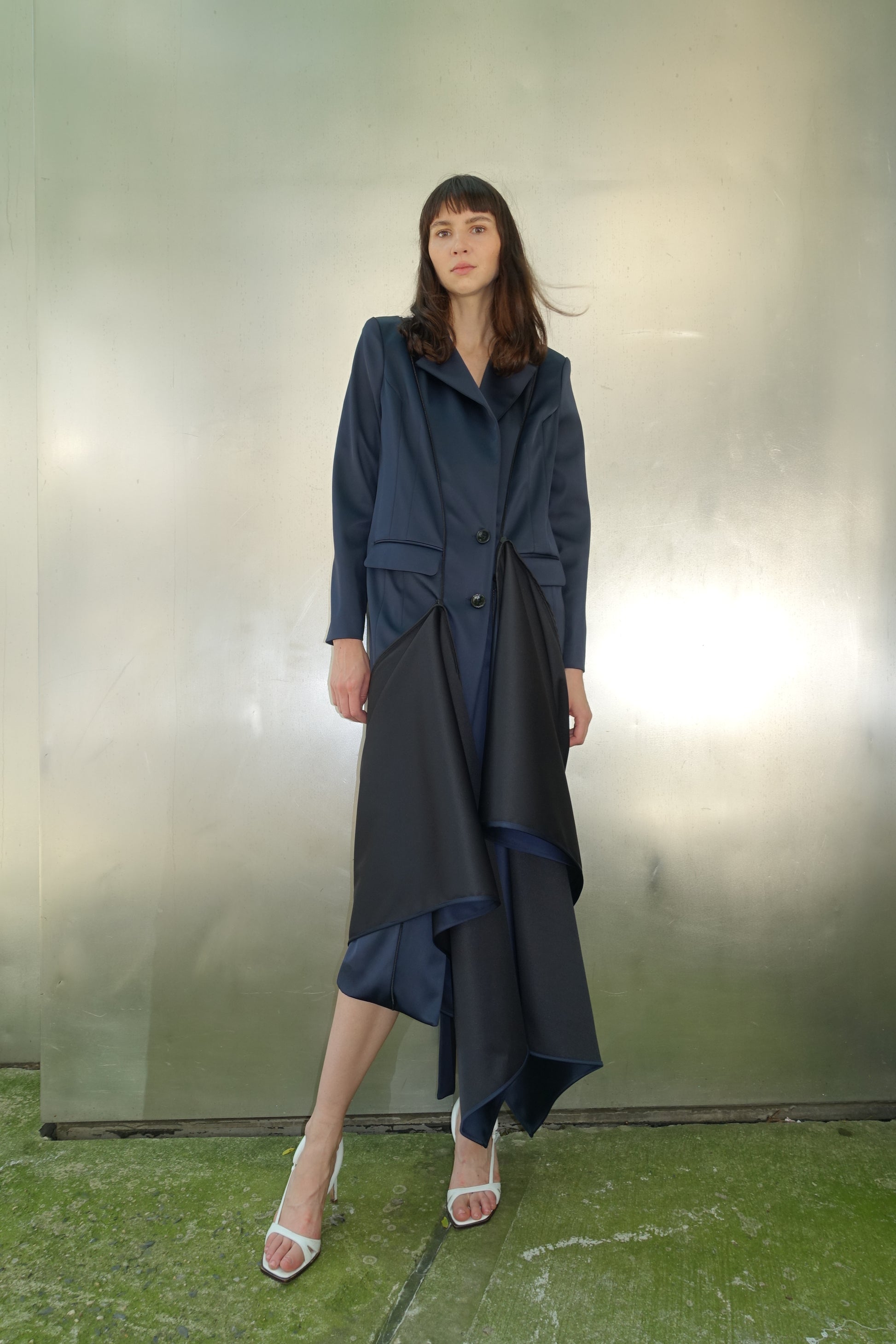 Woman wearing navy navy stretch satin coat with transformable zippers and self tie belt with draped detail on the lower side of her body