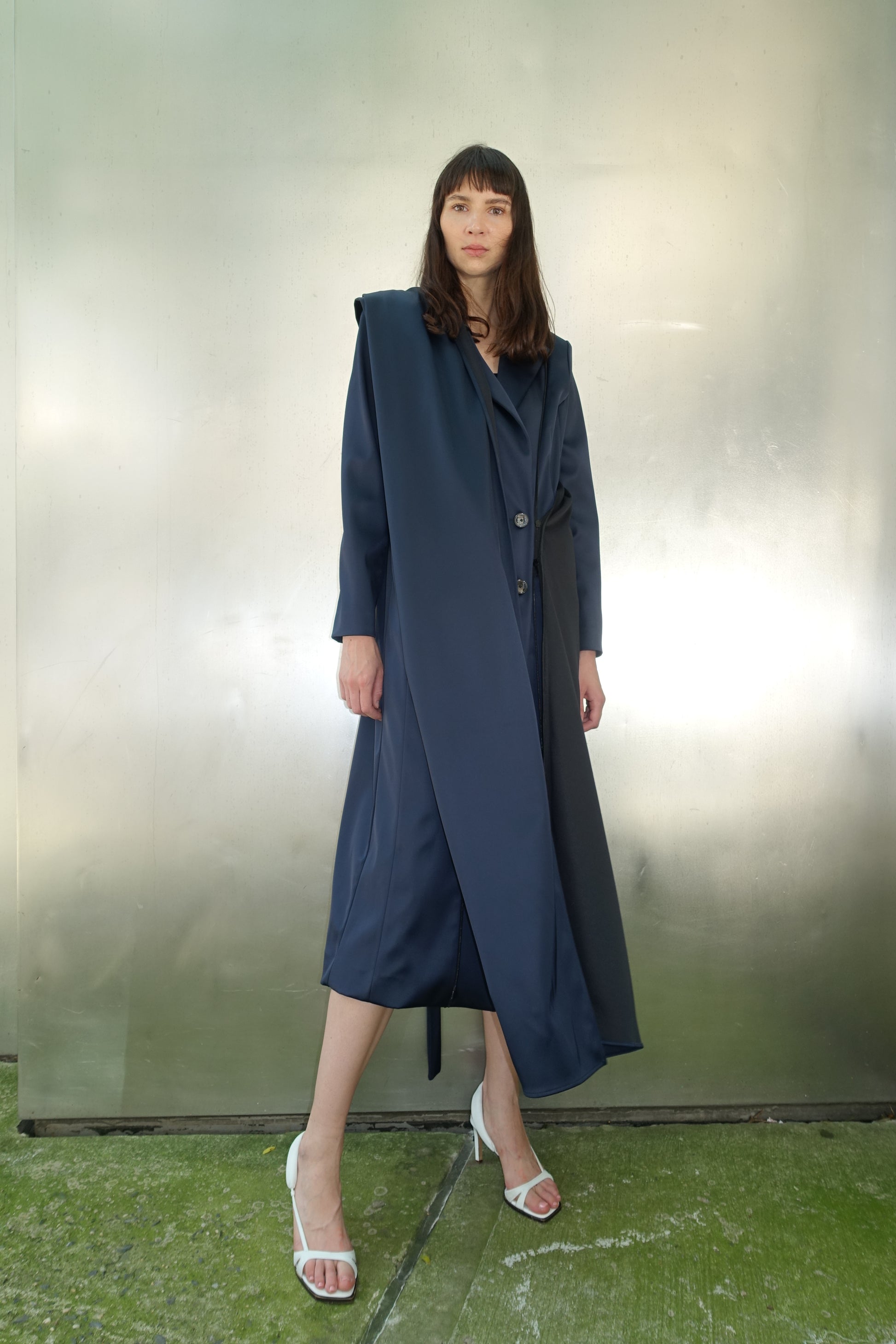 Woman wearing navy navy stretch satin coat with transformable zippers and self tie belt with draped detail on the right side of her body