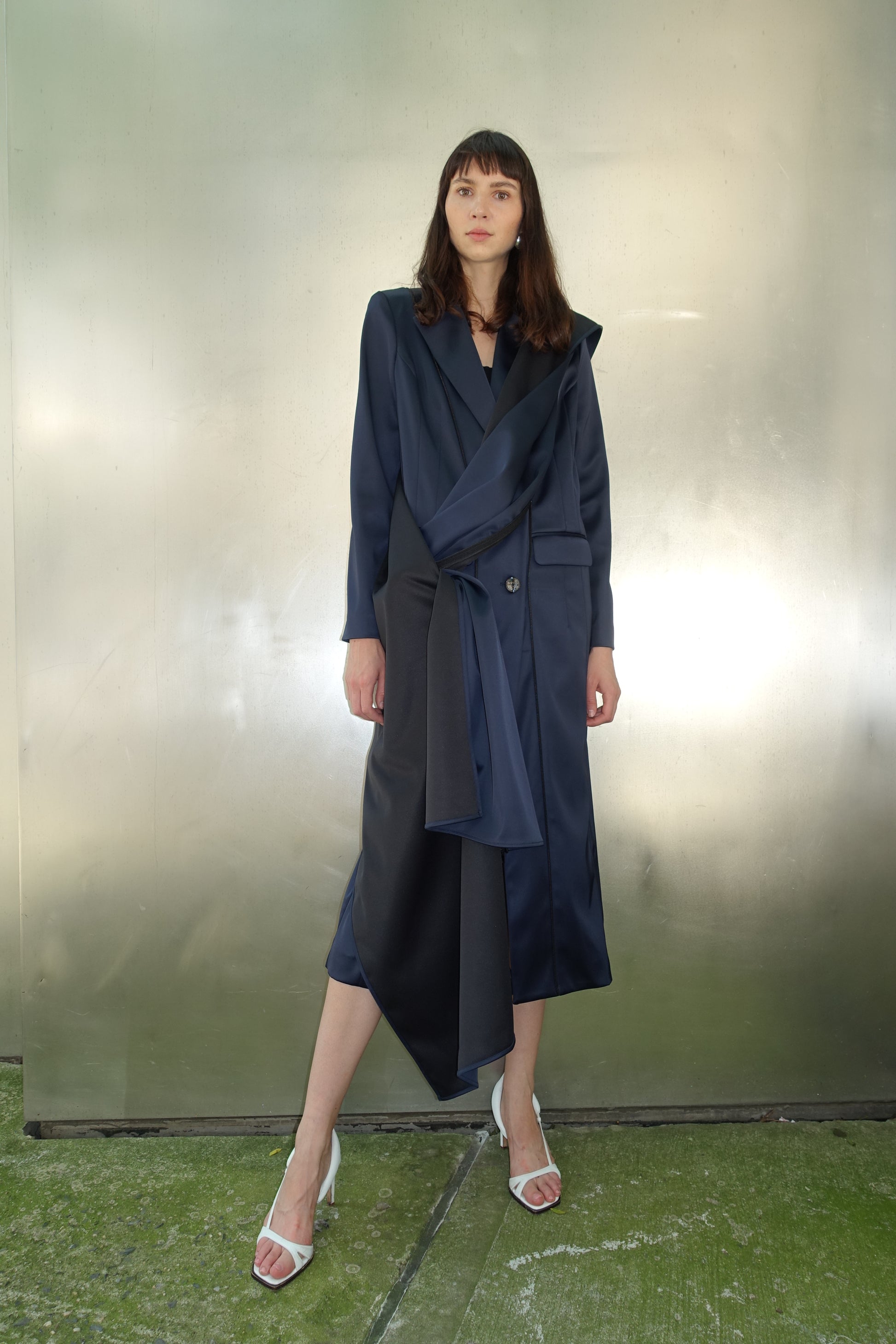 Woman wearing navy navy stretch satin coat with transformable zippers and self tie belt with draped detail around the waist 