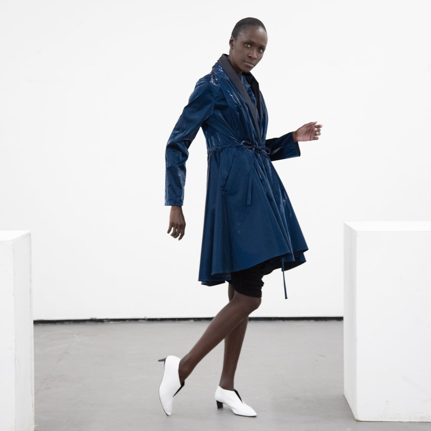 Woman in a blue shiny cotton coat with self tie belt and black detailing with white heels