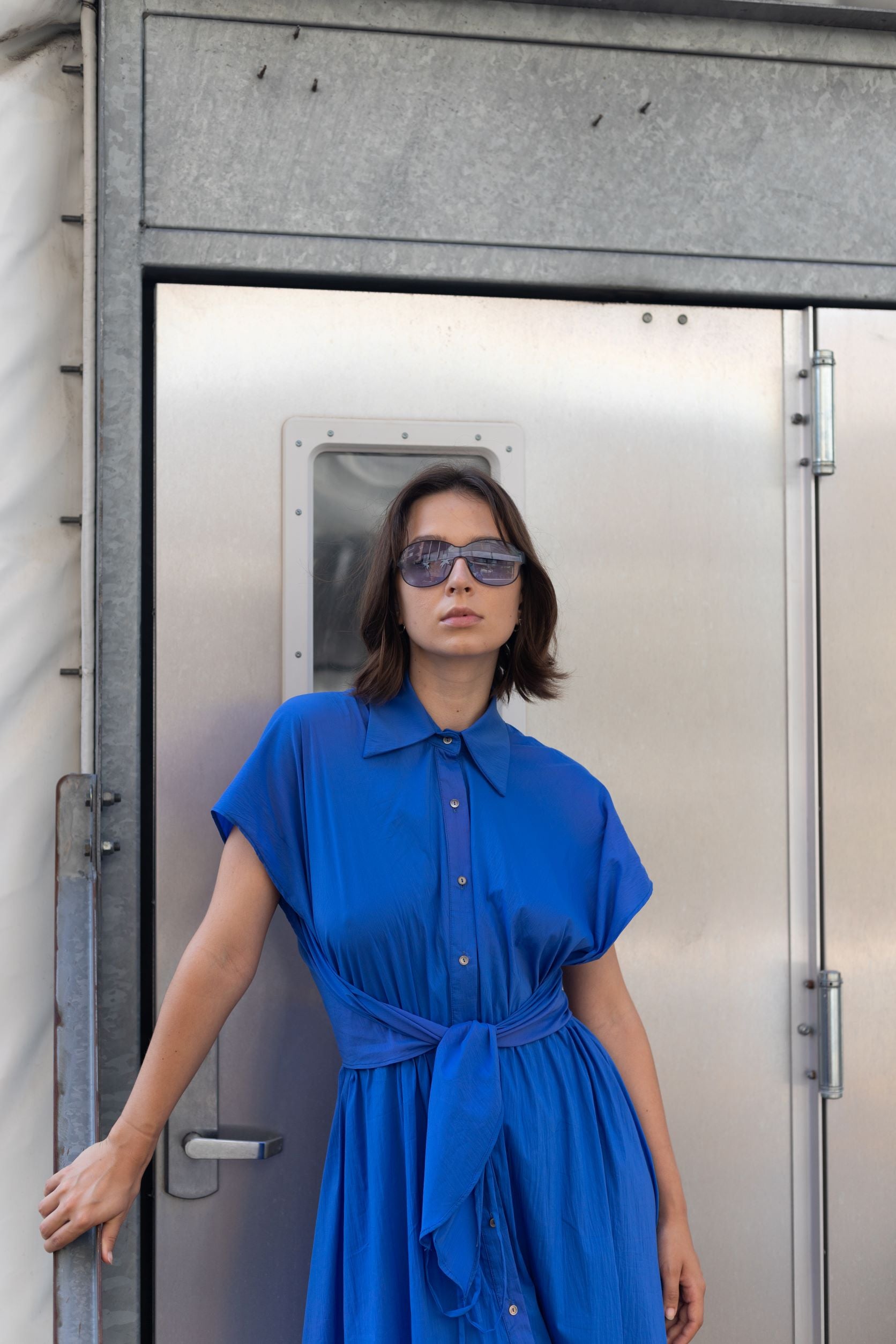 Woman wearing a cobalt blue cupro dress with button detailing and adjustable waist styled as belt and sunglasses