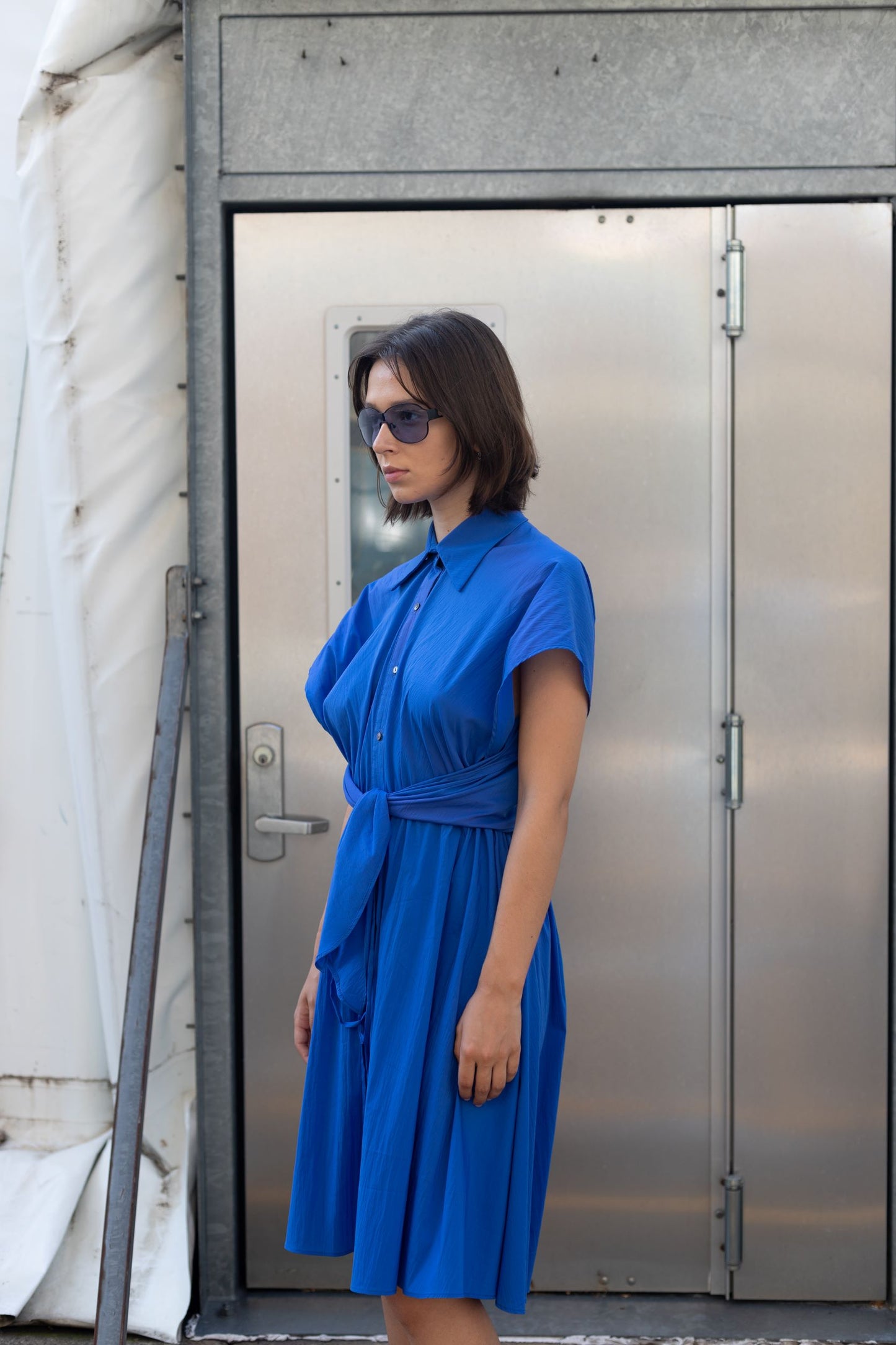 Woman in a cobalt blue cupro dress with button detailing and adjustable waist styled as belt and sunglasses
