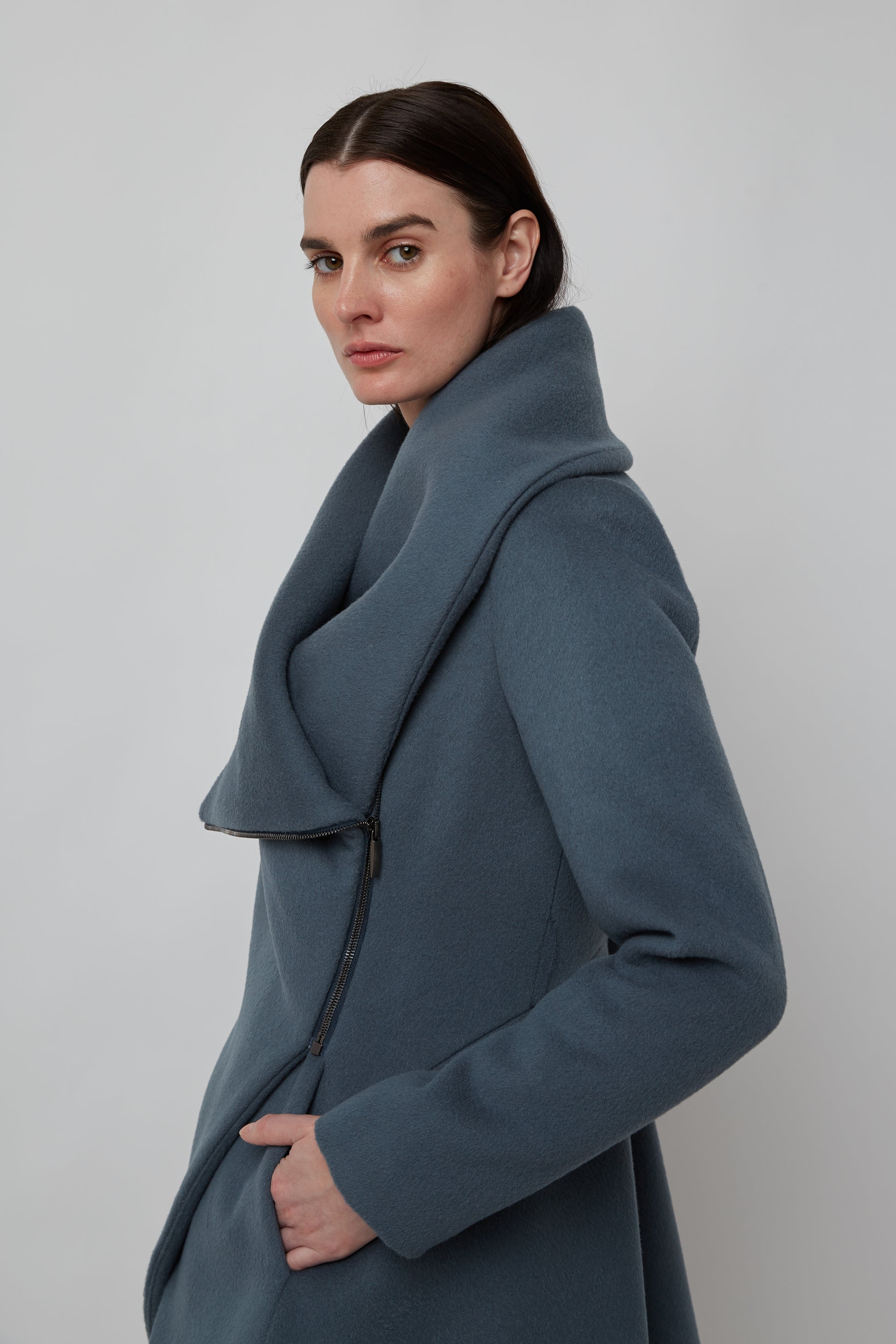 Woman wearing blue navy sculptural coat with asymmetric closure and hem line, with front zipper facing the side